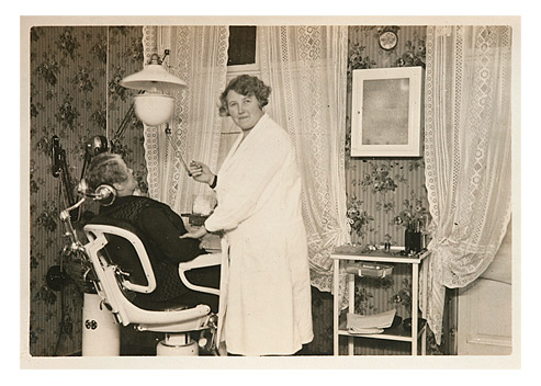 Eine freundliche Frau im Arztkittel steht vor einem Zahnarztstuhl, auf dem bereits eine Patientin sitzt. Der Raum erinnert mit seiner Blumentapete und den Rüschenvorhängen eher an ein bürgerliches Wohnzimmer als an eine Zahnarztpraxis.