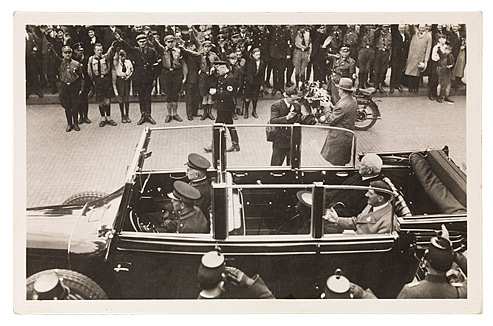 Schwarzweißfoto im Querformat, das ein Auto mit offenem Verdeck zeigt, in dem Hindenburg und Hitler und uniformierte Begleiter sitzen. Im Hintergrund Menschen in Zivil und Uniform, die Arme zum Hitlergruß erhoben. An der rechten Seite des Autos sind zwei Fotografen zu sehen