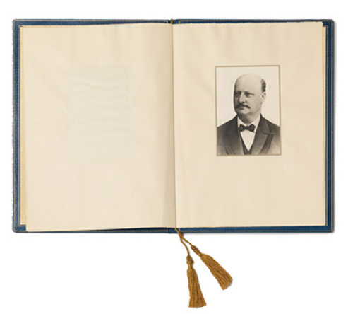 Aufgeschlagenes Buch, auf der rechten Seite eine Porträtfotografie eines schnurbärtigen Mannes in dunklem Anzug mit Fliege