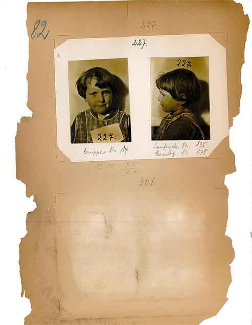Vergilbtes, an den Rändern angefressenes Blatt Papier, auf dem zwei Fotografien eines kleinen Mädchens aufgeklebt sind