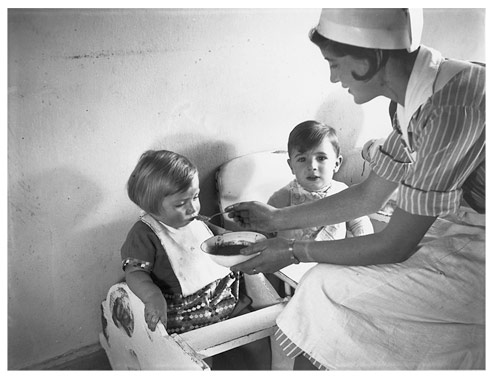 Eine Pflegerin füttert zwei Kleinkinder in Kinderstühlen.