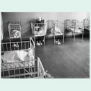 Raum mit mehreren Gitterbettchen, in denen Säuglinge liegen. Eine Pflegerin beugt sich über eines der Bettchen.