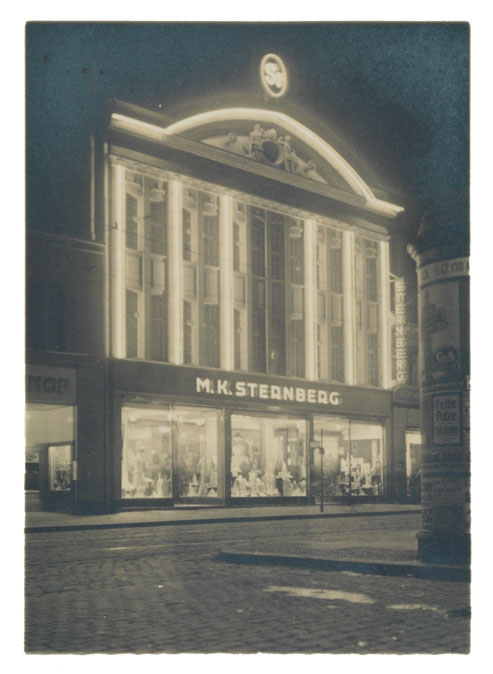 Hochformatige Fotografie, die ein stattliches Kaufhausgebäude bei Nacht mit Leuchtreklame und großen beleuchteten Schaufenstern zeigt.