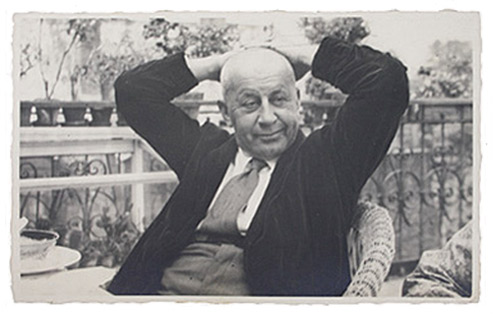 Portrait photo of an older man sitting in a wicker chair with his arms folded behind his head.