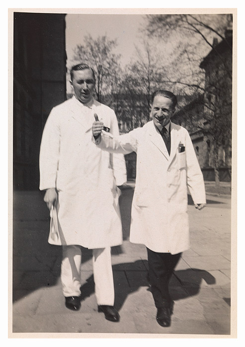 Two men in white lab coats crossing a square with the hospital in the background.