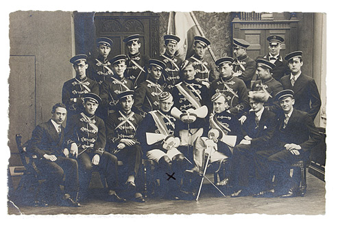 Twenty young members of a fraternity in a group photo. The three students with leading positions are dressed in Vollwichs (full-dress uniform) and are holding their swords. The other members are wearing Kneipjacken—i.e., suits, caps and colored bands.