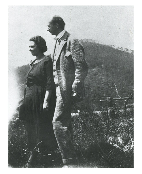 A couple taking a walk. The woman wears a dark summer dress, while the tall man is clad in a suit and carries a walking stick. They are walking through long grass with a hill in the background.