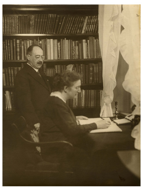 A woman sits at a desk writing something down. Behind her stands a man in front of a bookshelf.