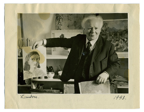 An elderly man in a dark suit with a fringe of white hair and a friendly face. He is presenting a watercolor drawing of a woman‘s head and there are shelves full of art supplies and paintings in the room behind him.