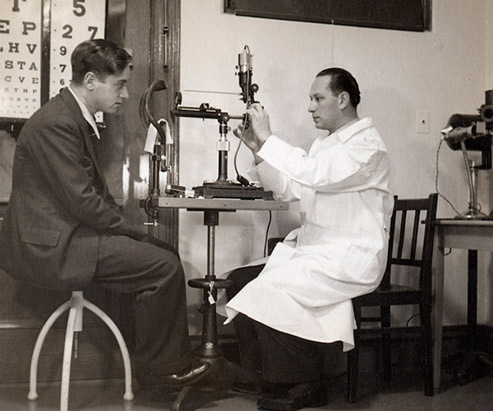 View of an eye doctor‘s office with a doctor in a white coat sitting on the right adjusting an examination instrument. A patient is sitting in front of him.