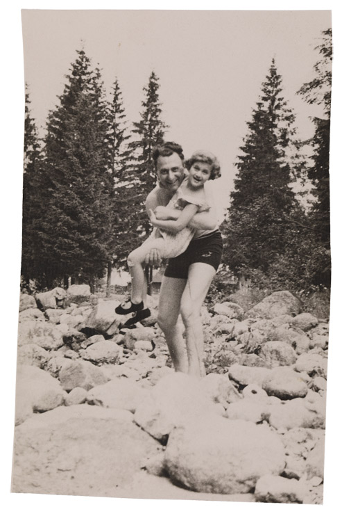 Vertical-format photo showing a man in a bathing suit with a girl in his arms wearing a summer dress. They are standing amidst large stones, perhaps in a dry river bed, and looking cheerfully at the camera. Fir trees can be seen in the background.
