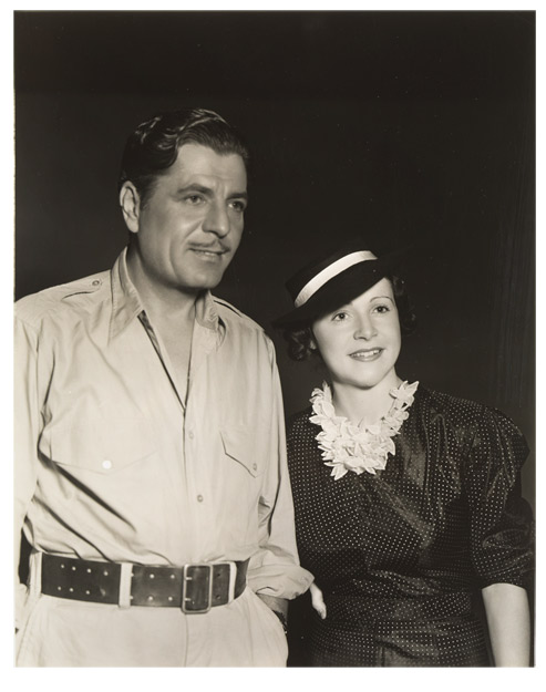 Photograph of a man with a thin mustache wearing an open-necked shirt, pants and a wide belt. Standing at his side is a young woman in a dark dress and hat.