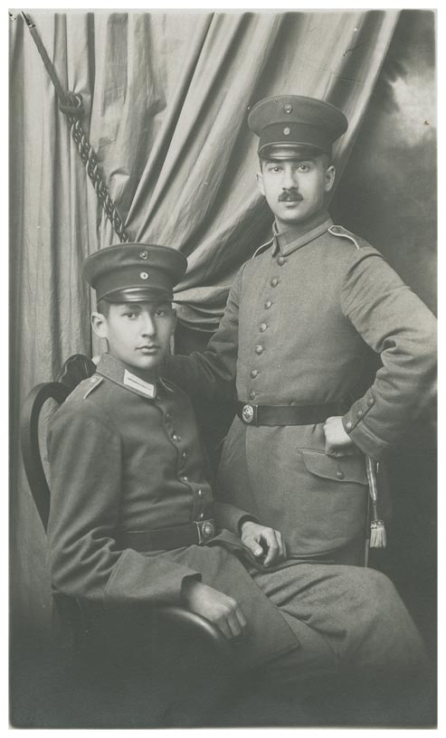 Two young men in uniform, one sitting on a chair, the other standing behind him. Judging by the carefully draped curtain, the picture was taken in a photography studio.