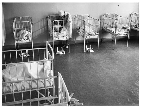 Room containing several cribs with infants. A nurse is leaning over one of the small beds.