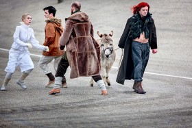 An old man and a young man, a person dressed in white and a man dressed in dark colours with long red hair and a donkey, all moving on tarmac