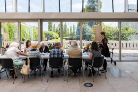 A coffee table with elderly people, behind it a window front offers a view of the garden of the Jewish Museum Berlin.