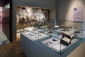 Blick in einen Ausstellungsraum, an dessen hinterer Wand der Gipsabguss des Titusbogens zu sehen ist, im Vordergrund eine Vitrine mit Menora, Tora-Aufsätzen und aufgeschlagenen Büchern