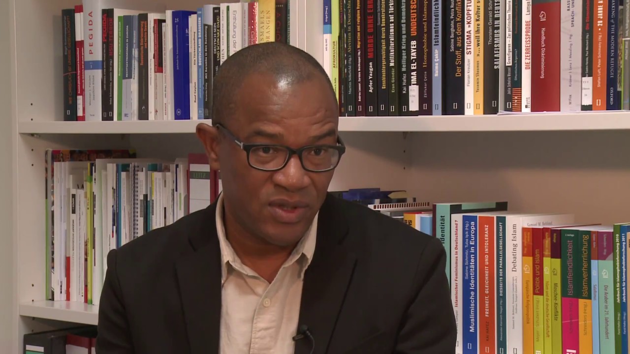 A man with glasses is sitting in front of a bookshelf giving an interview.