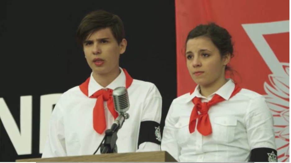 Zwei junge Menschen in weißen Hemden und Roten Halstüchern stehen auf einer Bühne.