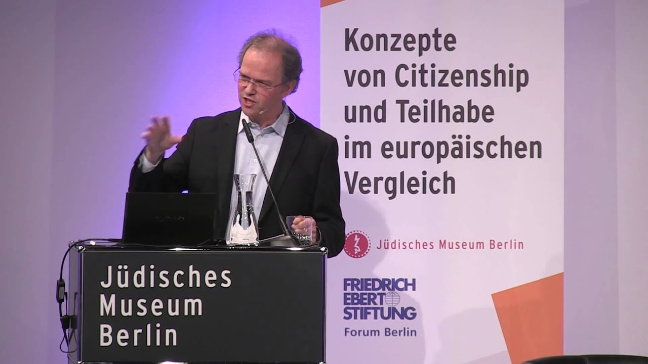 Man with suit and glasses stands at lectern and gives a lecture.