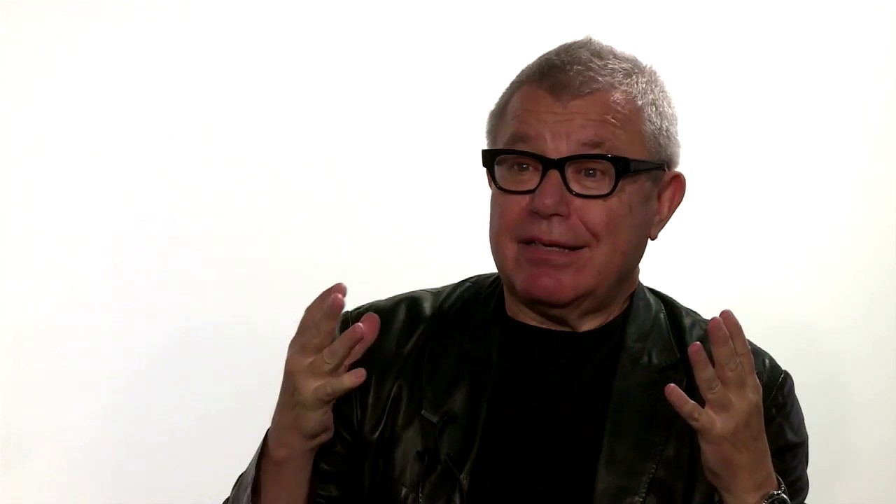 Man with gray hair and glasses gestures with both hands.