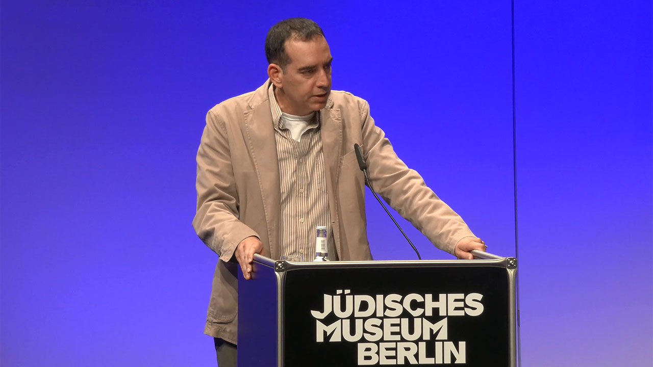 Man in beige blazer stands at a lectern.