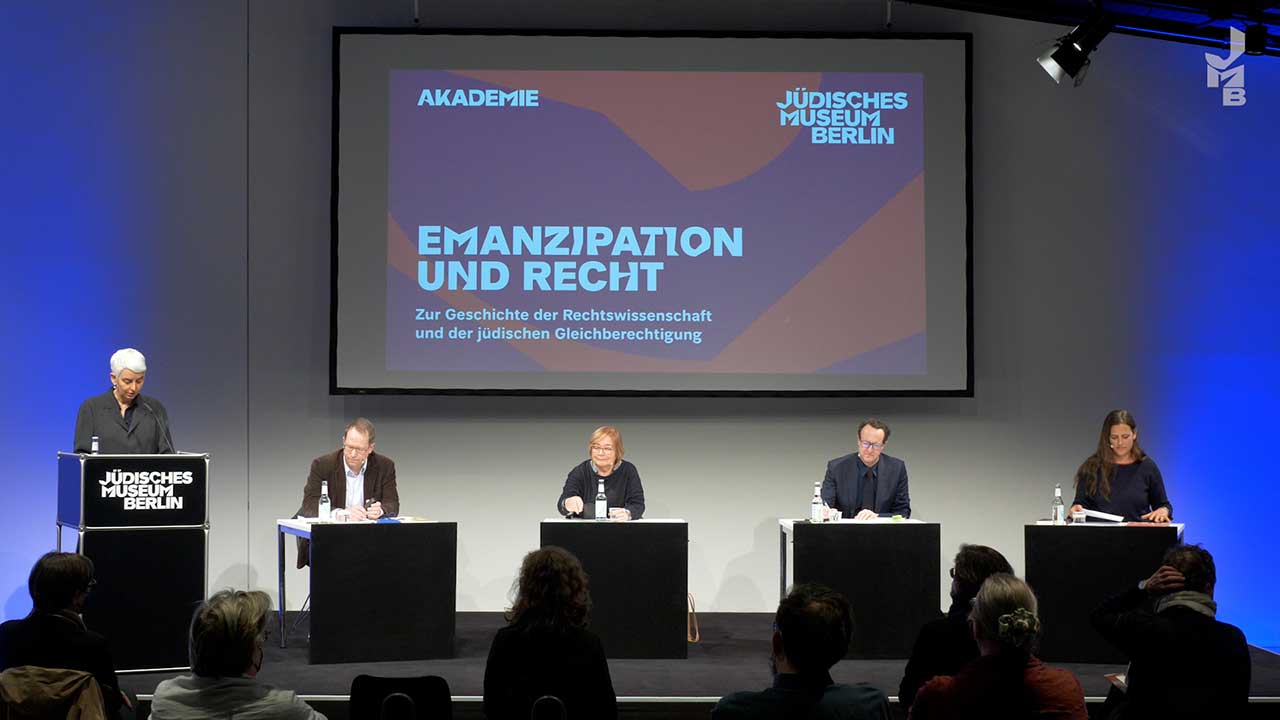 Stage on which a woman stands at a lectern and four people sit at small tables.