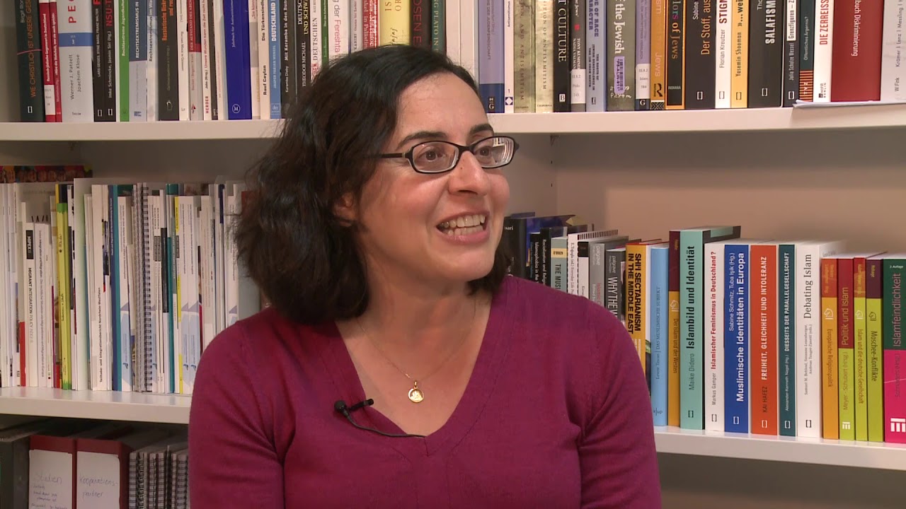 A woman with a red sweater is sitting in front of a bookshelf giving an interview.