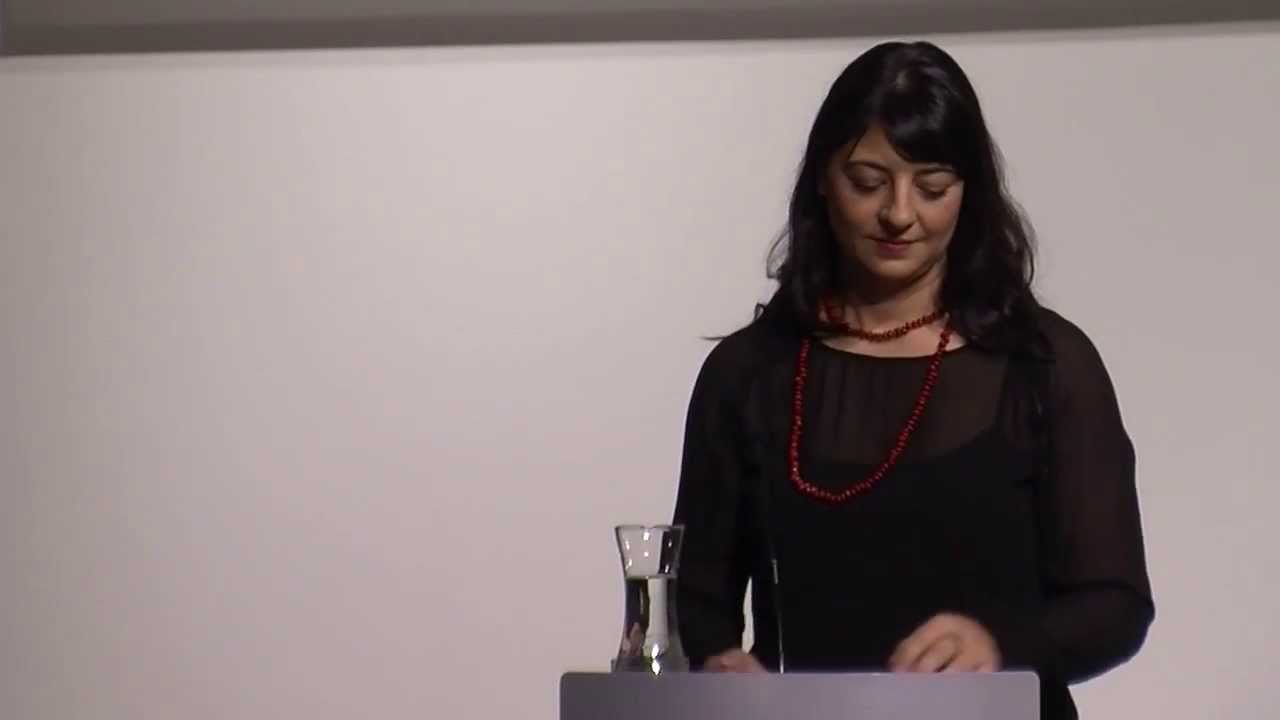 A woman stands at a lectern and reads something aloud.