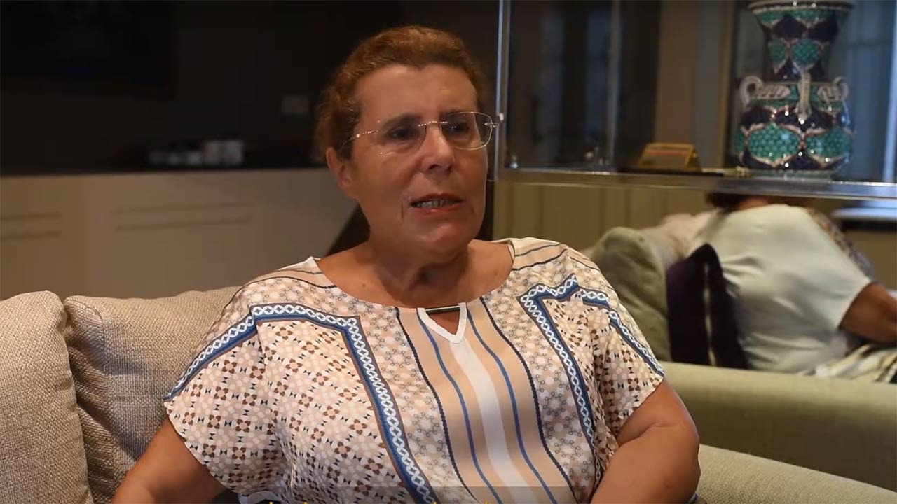 Video still: A woman with glasses sits on a sofa and speaks.