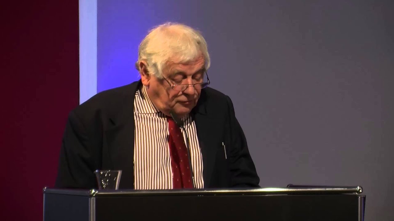 Man with white hair stands at lectern and speaks.