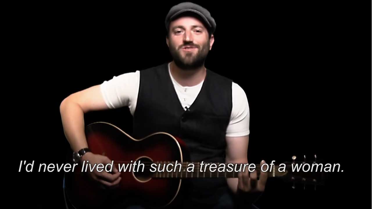 Daniel Kahn mit Gitarre vor schwarzem Hintergrund. Folgende Untertitel sind zu sehen: I've never lived with such a treasure of a woman.