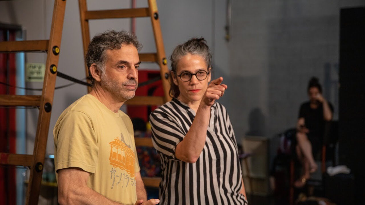 A woman and a man are standing next to each other in a room. A stepladder can be seen behind them. The woman points in the direction next to the camera. The man's thoughts seem to be elsewhere.