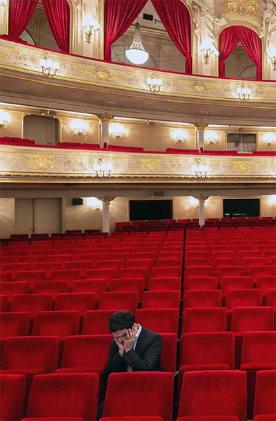 Fotografie einer Person allein auf einem der roten Sitze im Parkett eines leeren Theaters, den Kopf in die Hände gestützt
