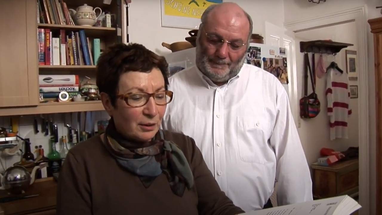 Video still: A woman with short brown hair and glasses shows a book to a bearded man in a white shirt.