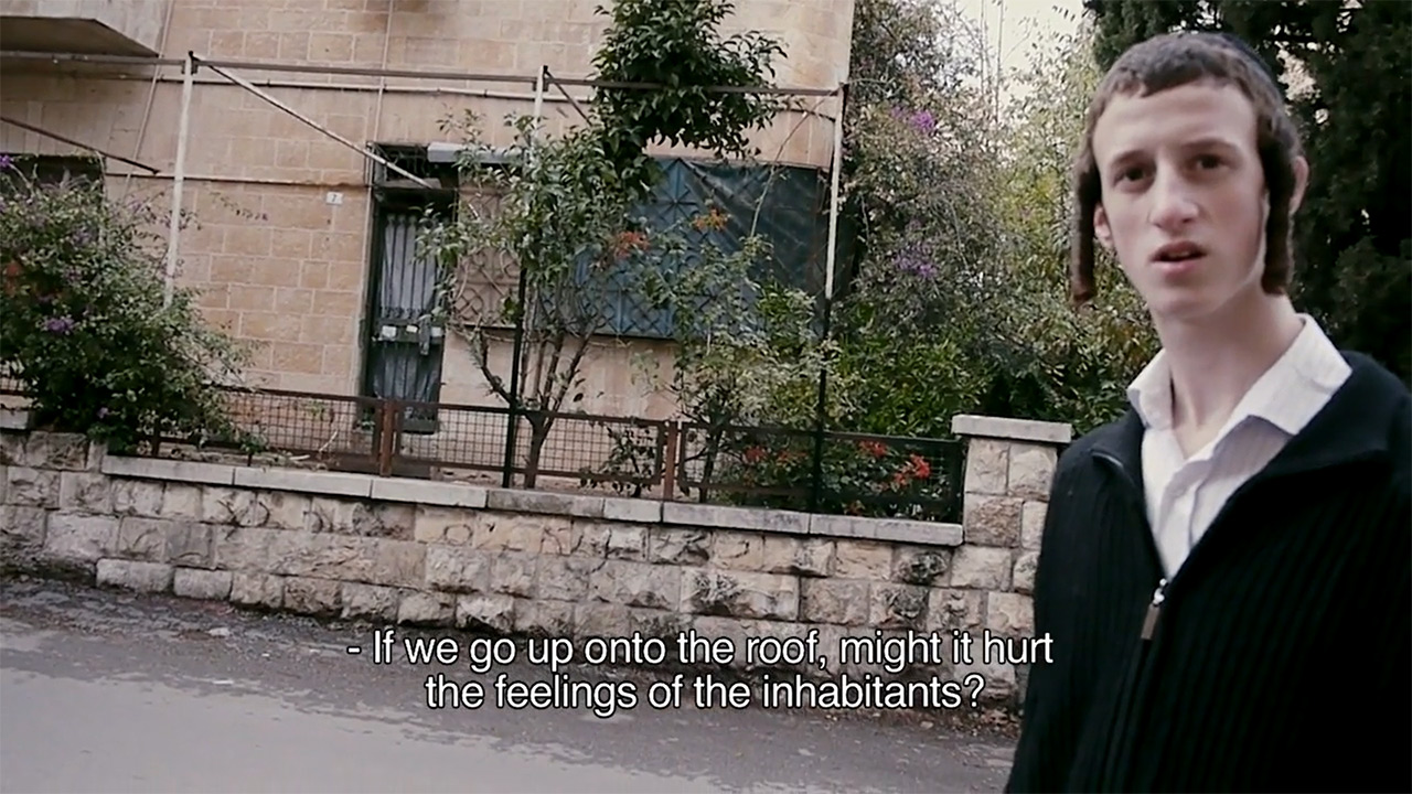 Young man with a kippah and temple curls stands on a street and looks into the camera.