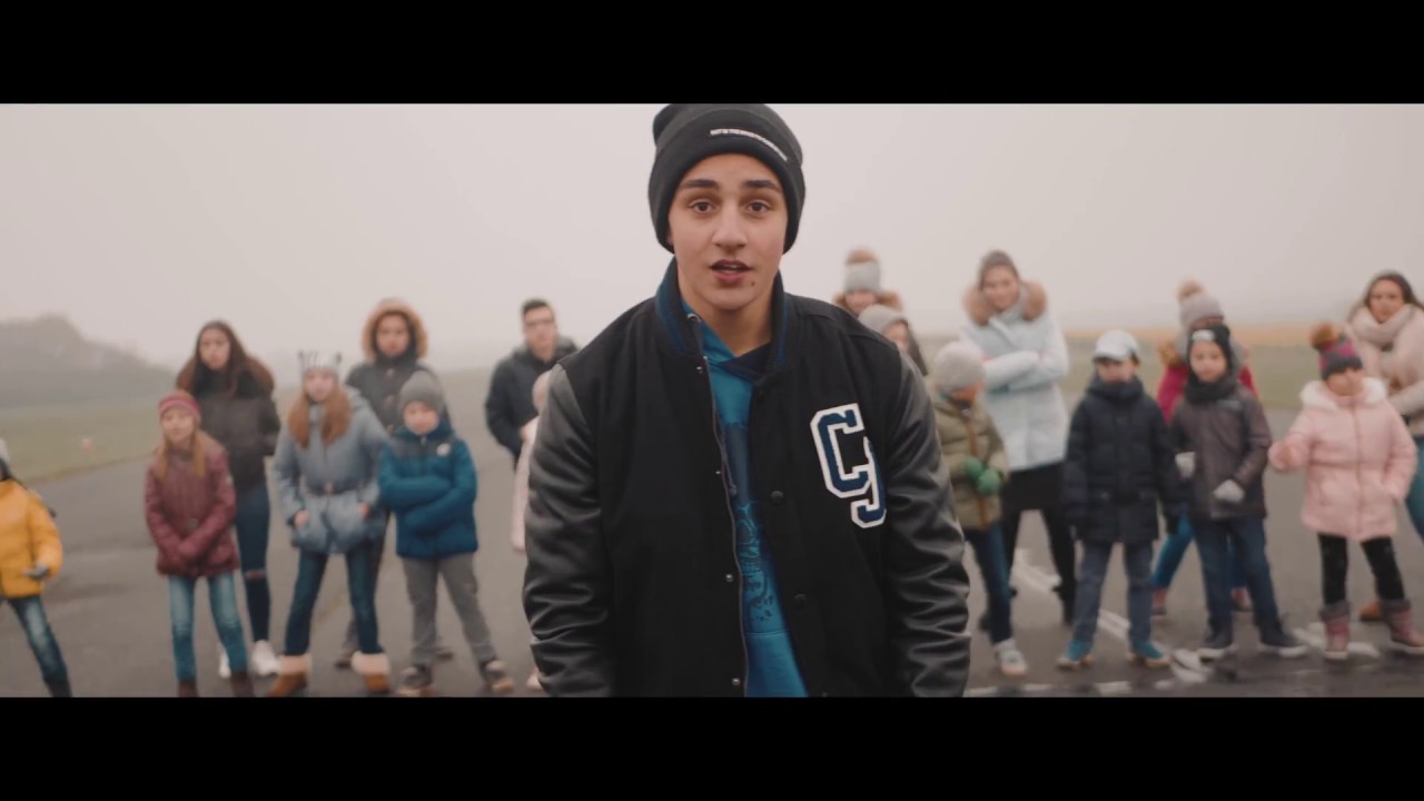 Young person with cap looks at camera and sings. In the background there is a group of young people dancing.