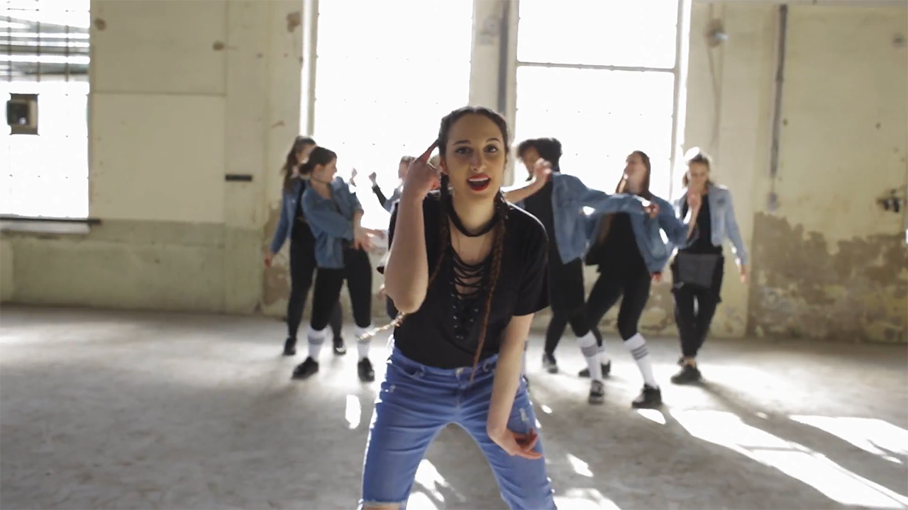 Young woman dancing and singing in a sunlit room with other dancers.