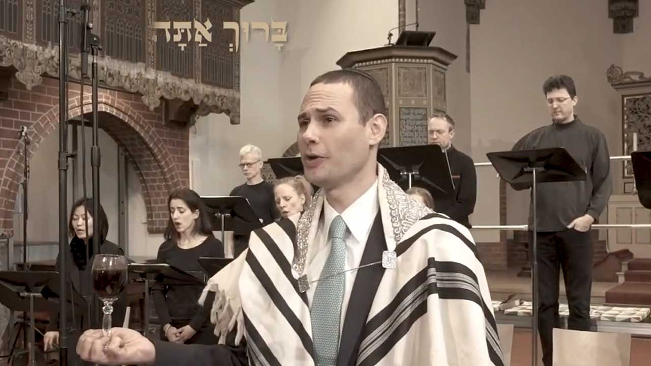 Cantor Azi Schwartz sings the Kiddush. Other singers in black outfits can be seen in the background.