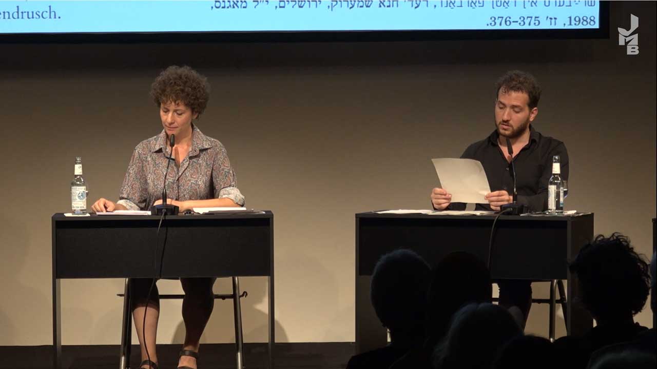 A woman and a man each sit at a table on a stage.
