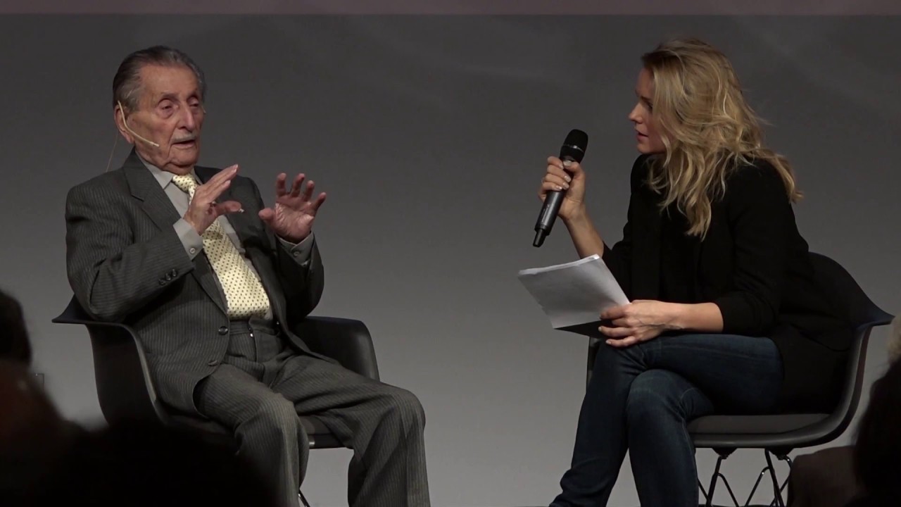 An older gentleman and a younger lady sitting on a podium and talking