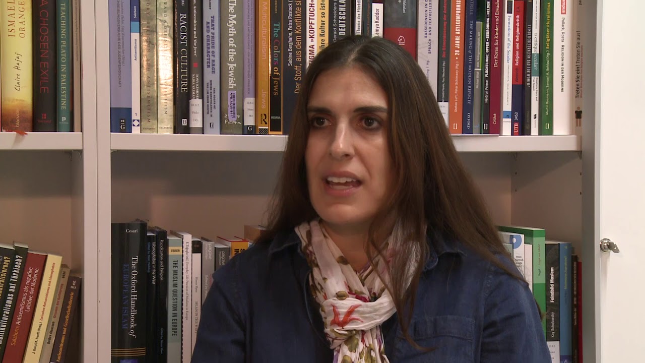 Woman with white hair and black jacket sits in front of a bookshelf and gives an interview.