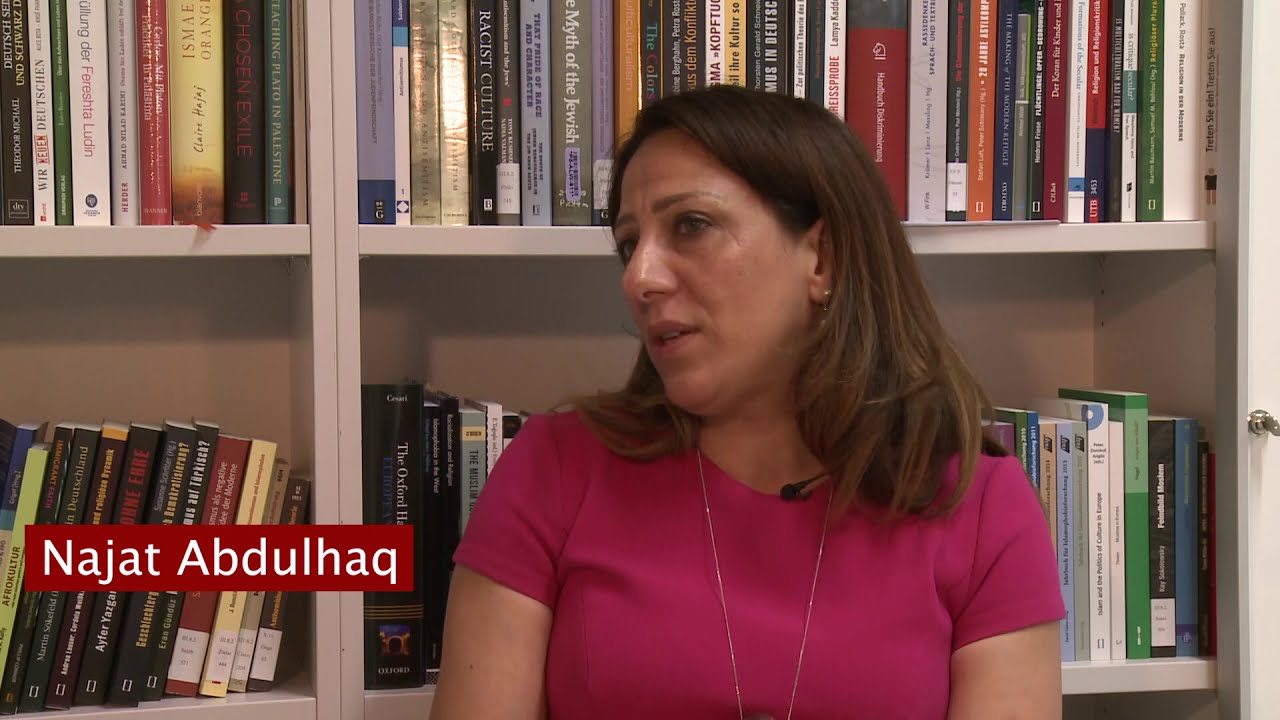 Woman in a pink t-shirt sits in front of a bookshelf giving an interview.