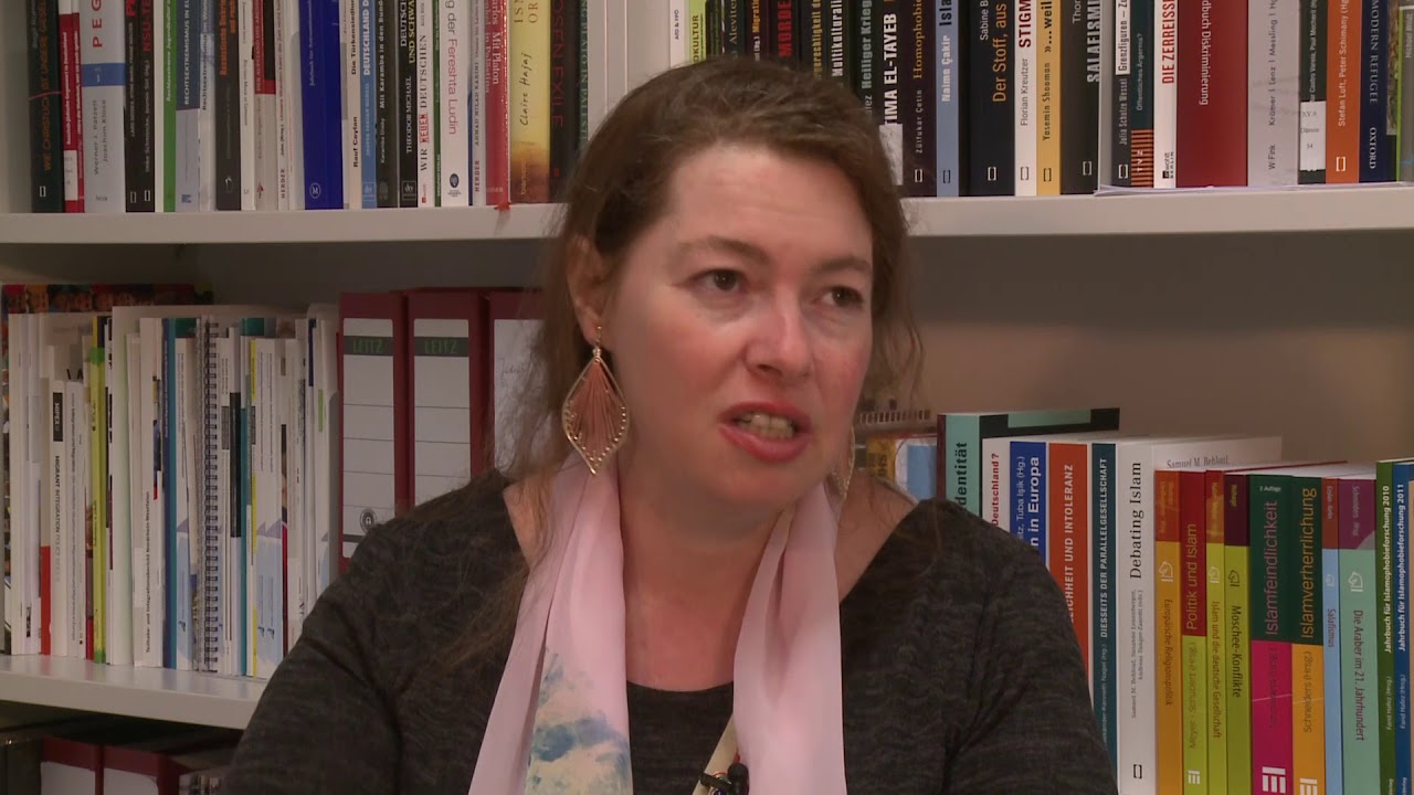 Woman with pink scarf sits in front of a bookshelf and gives an interview.