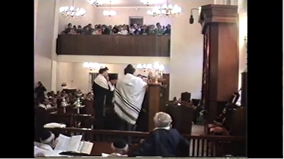 View into a synagogue filled with people, the rabbi is standing at the lectern.