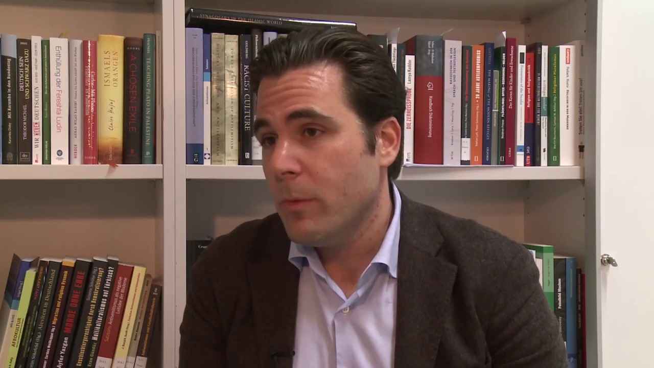 Man with jacket and shirt sits in front of a bookshelf and gives an interview.