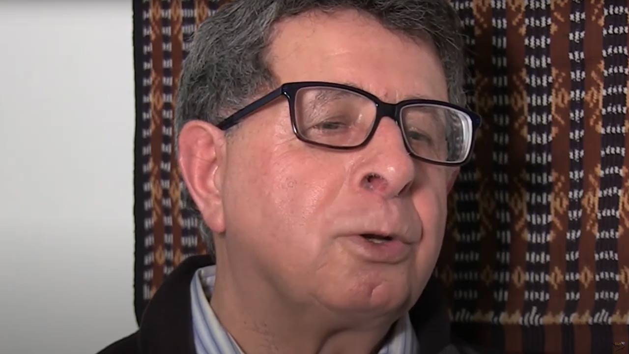 Video still: Face of a man with glasses in front of a wall with carpet hangings.