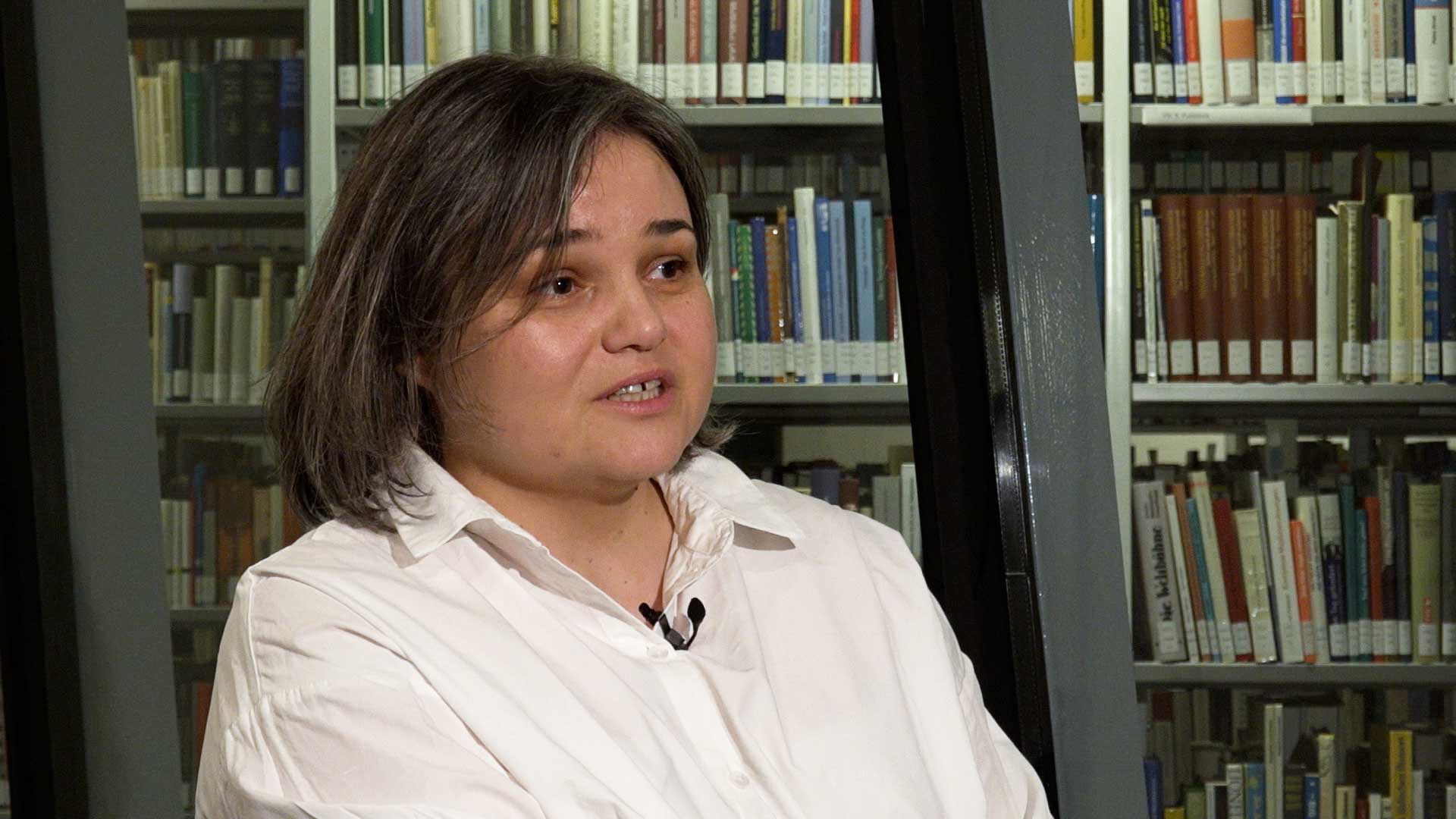 Woman with gray, chin-length hair speaks and sits in front of a window pane behind which bookshelves stand.