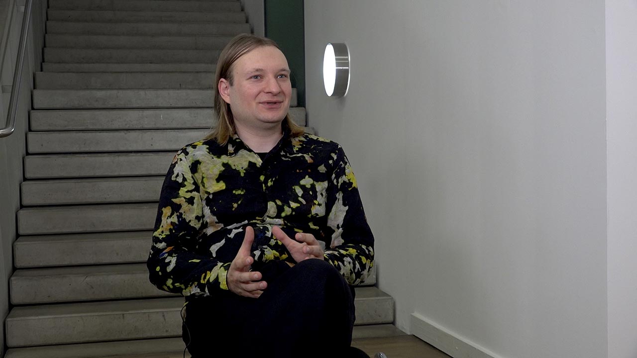 Ein junger Mann mit schulterlangem blondem Haar im geblümten Hemd sitzt gestikulierend vor einer Treppe.