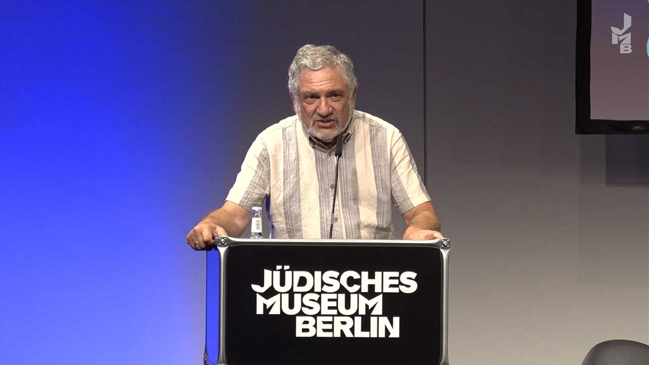 Mann steht vor blauen Hintergrund an einem Rednerpult, auf dem Jüdisches Museum Berlin steht.